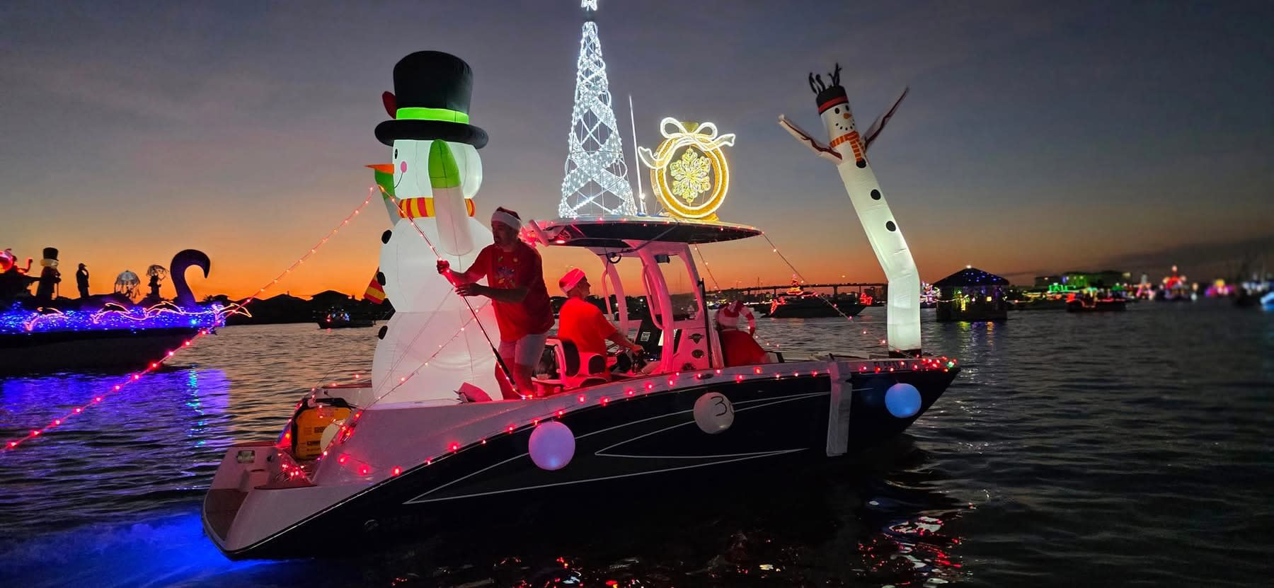 Sunset parade: boat with top-hat snowman and Santa, lit tree and wreath behind