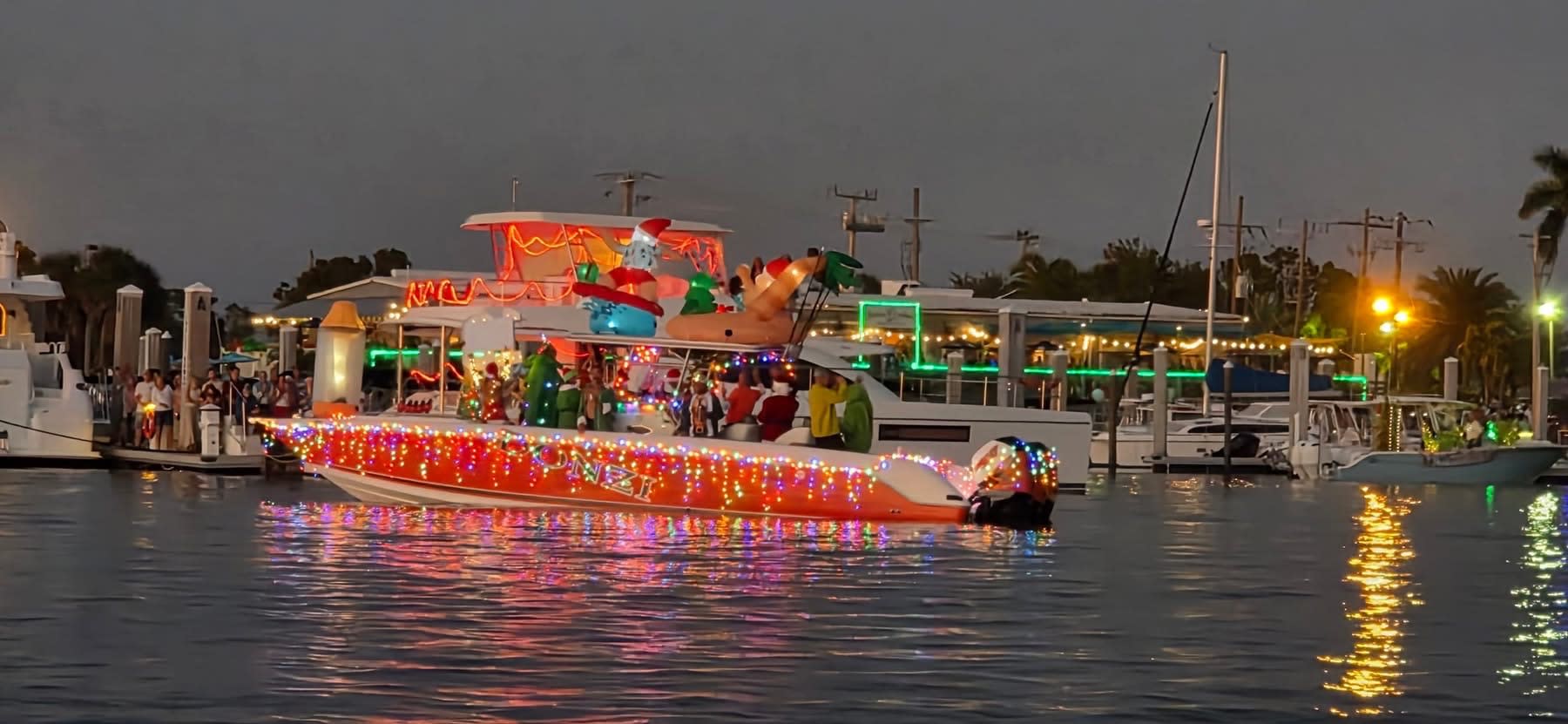 Large deck boat outlined in orange and multicolor lights, crowds aboard at dock