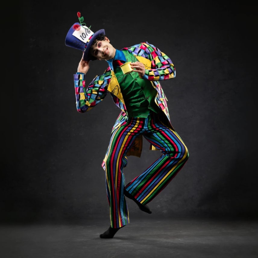 A dancer as the Mad Hatter with colorful striped pants and checkered suit jacket holding a cup of tea and tipping his top hat