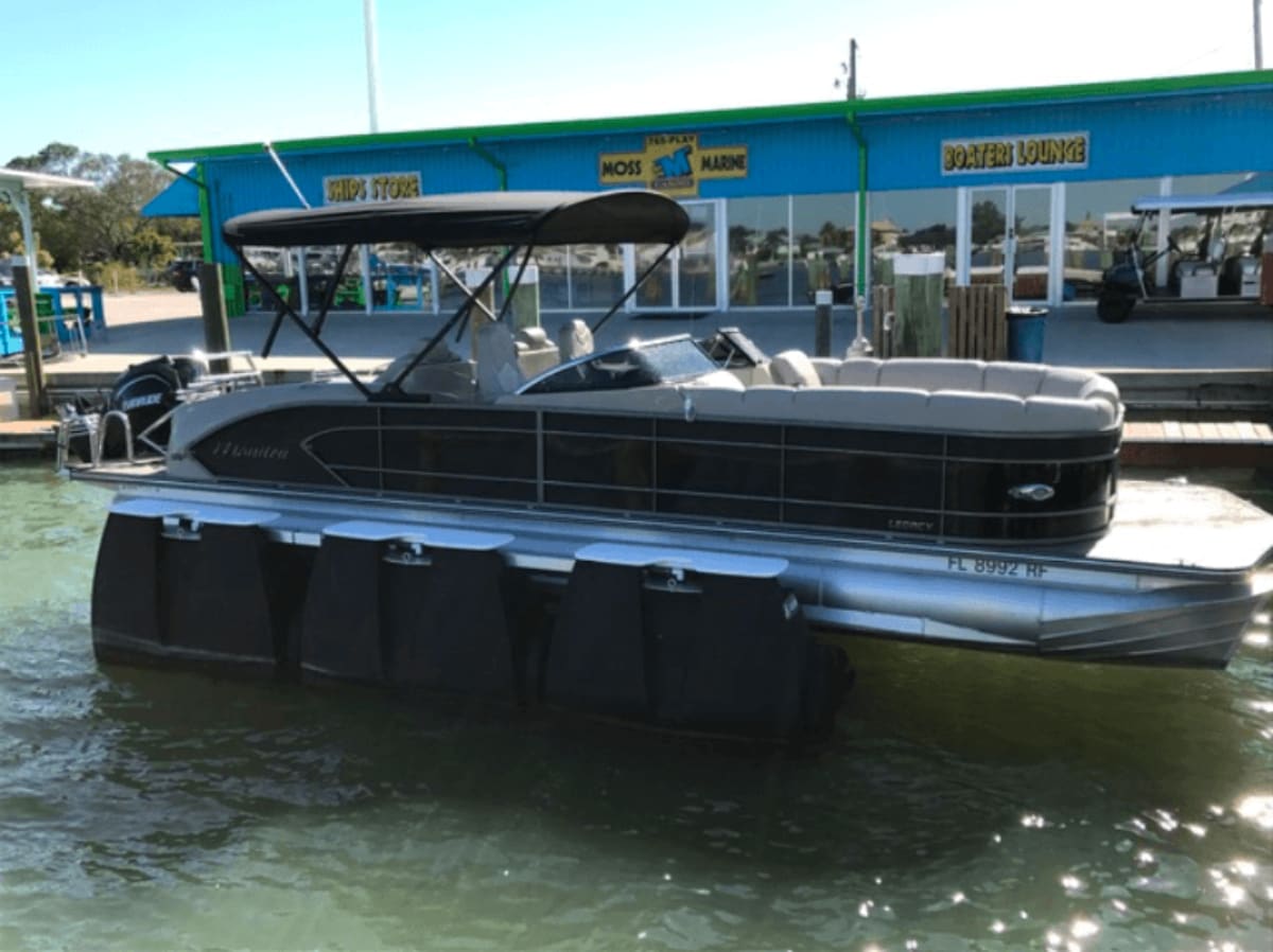 Black pontoon boat docked at Moss Marine facility with blue building and boat lounge signage visible.