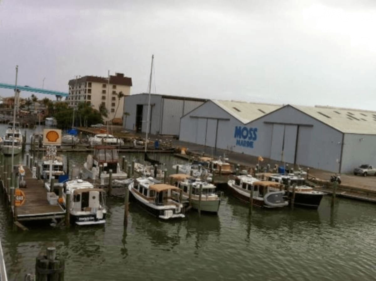 Harbor with multiple docked boats, Shell gas sign, and Moss Marine warehouse buildings along waterfront.