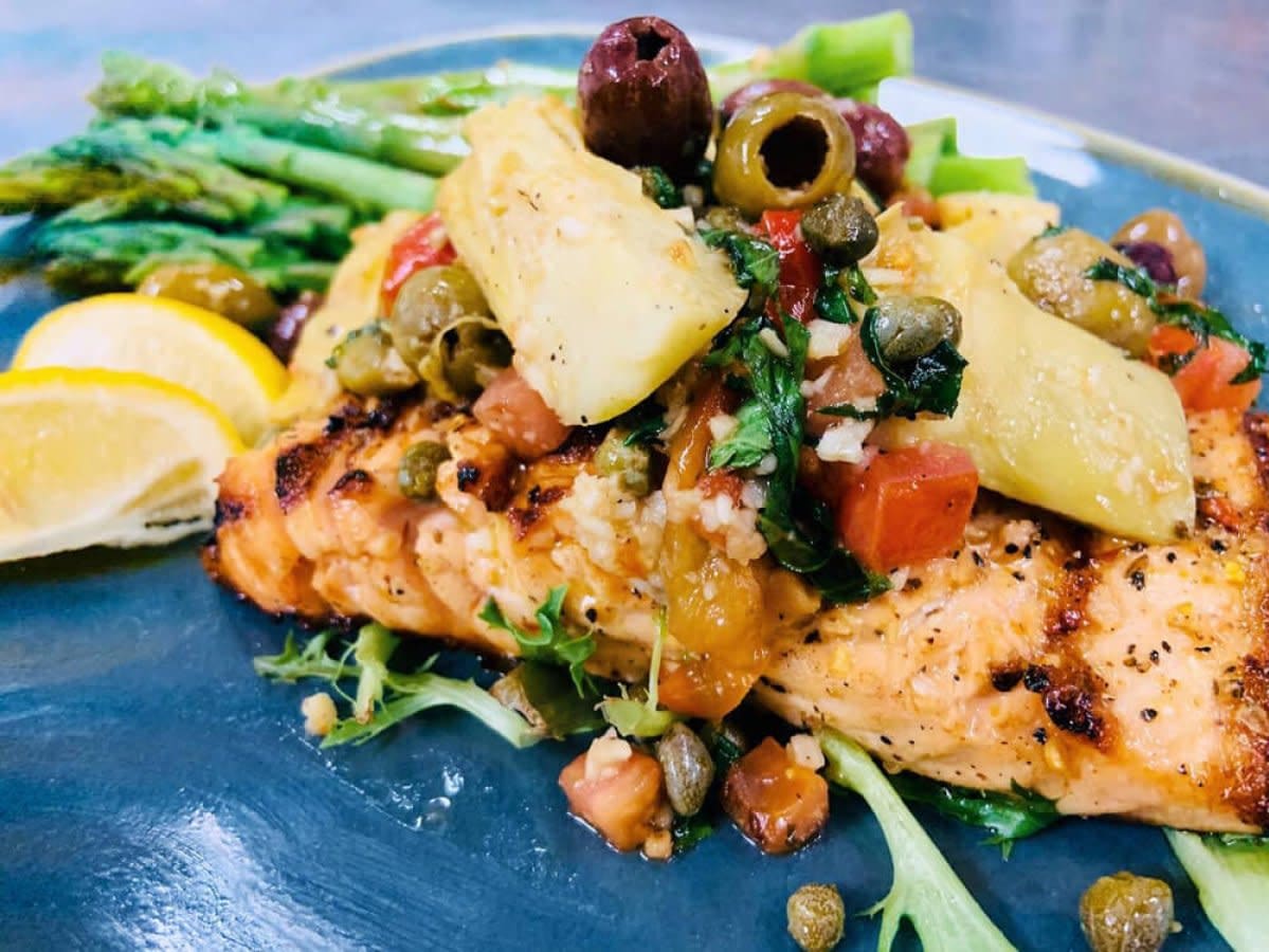 Grilled salmon plated with asparagus, pineapple, olives, capers, and lemon wedges