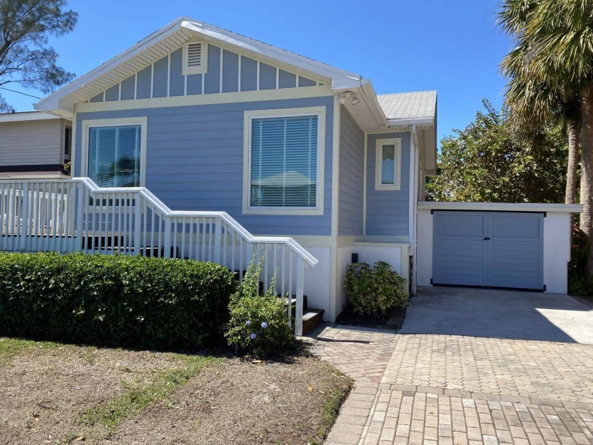 Two-story light blue beach house with white railings, brick driveway, garage door, palm trees, sunny day.