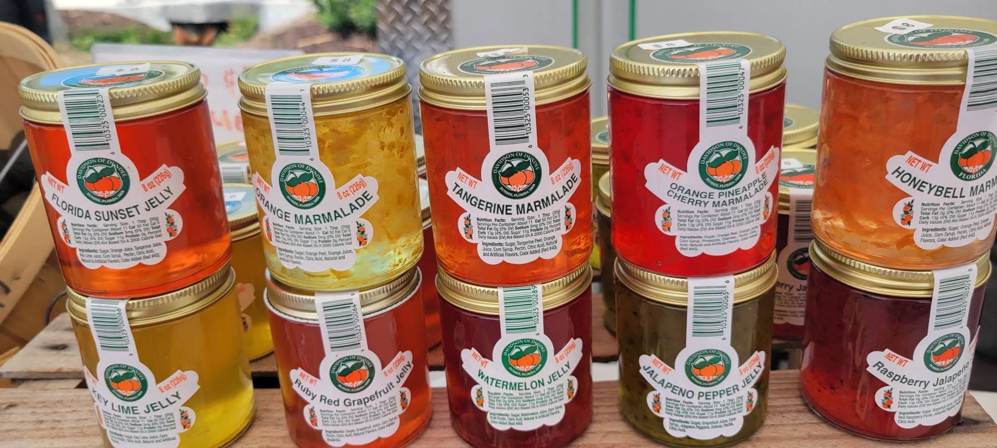 Stacked jars of colorful jams and marmalades including Florida Sunset, citrus flavors, and jalapeño varieties