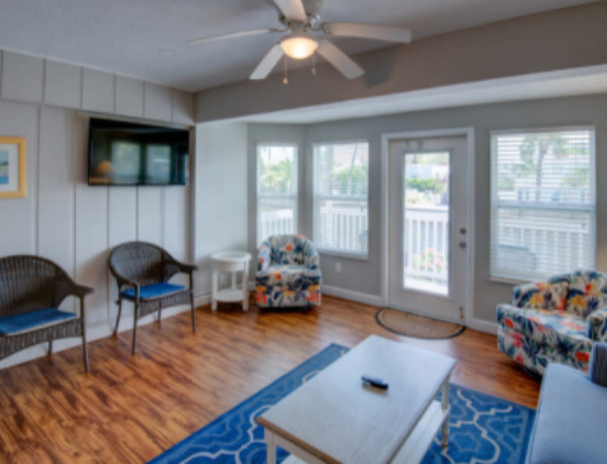 Bright living room with blue patterned rug, floral chairs, wall-mounted TV, and balcony access through glass doors.