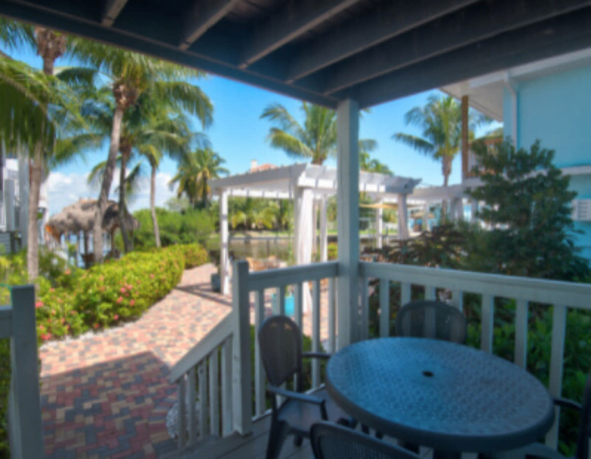Covered deck with round table and chairs overlooking tropical resort grounds with palm trees and brick pathway.