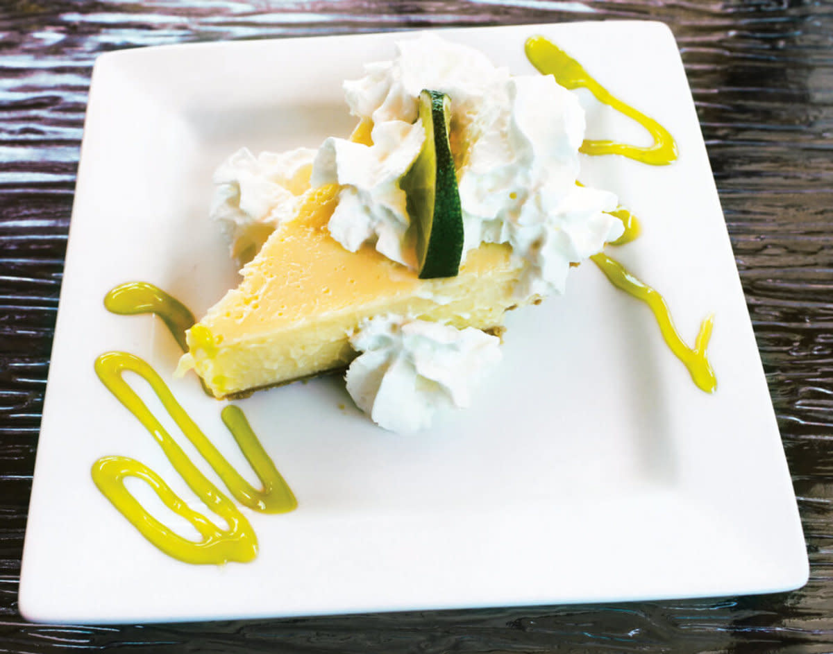 Lemon cheesecake slice on white plate topped with whipped cream, lime wedge, and yellow drizzle.
