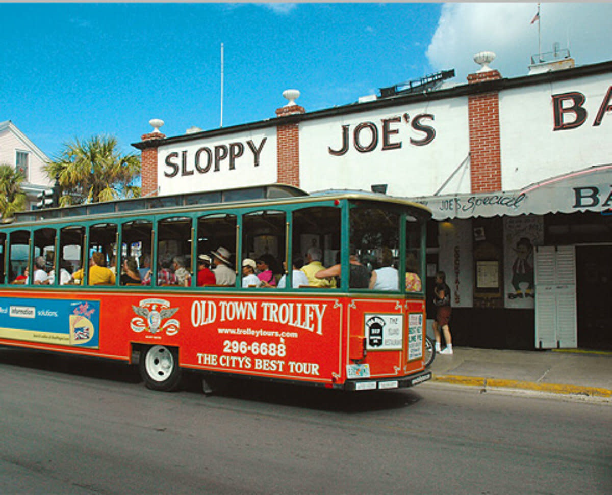Red Old Town Trolley bus passes historic Sloppy Joe's Bar building with tourists aboard on sunny street