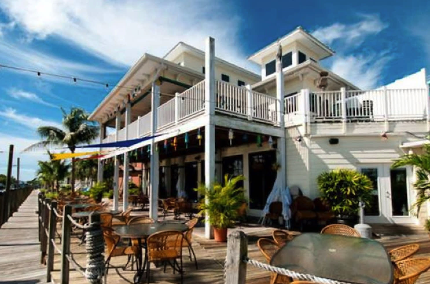 White waterfront restaurant with covered balcony, outdoor deck seating, palm trees, and blue sky
