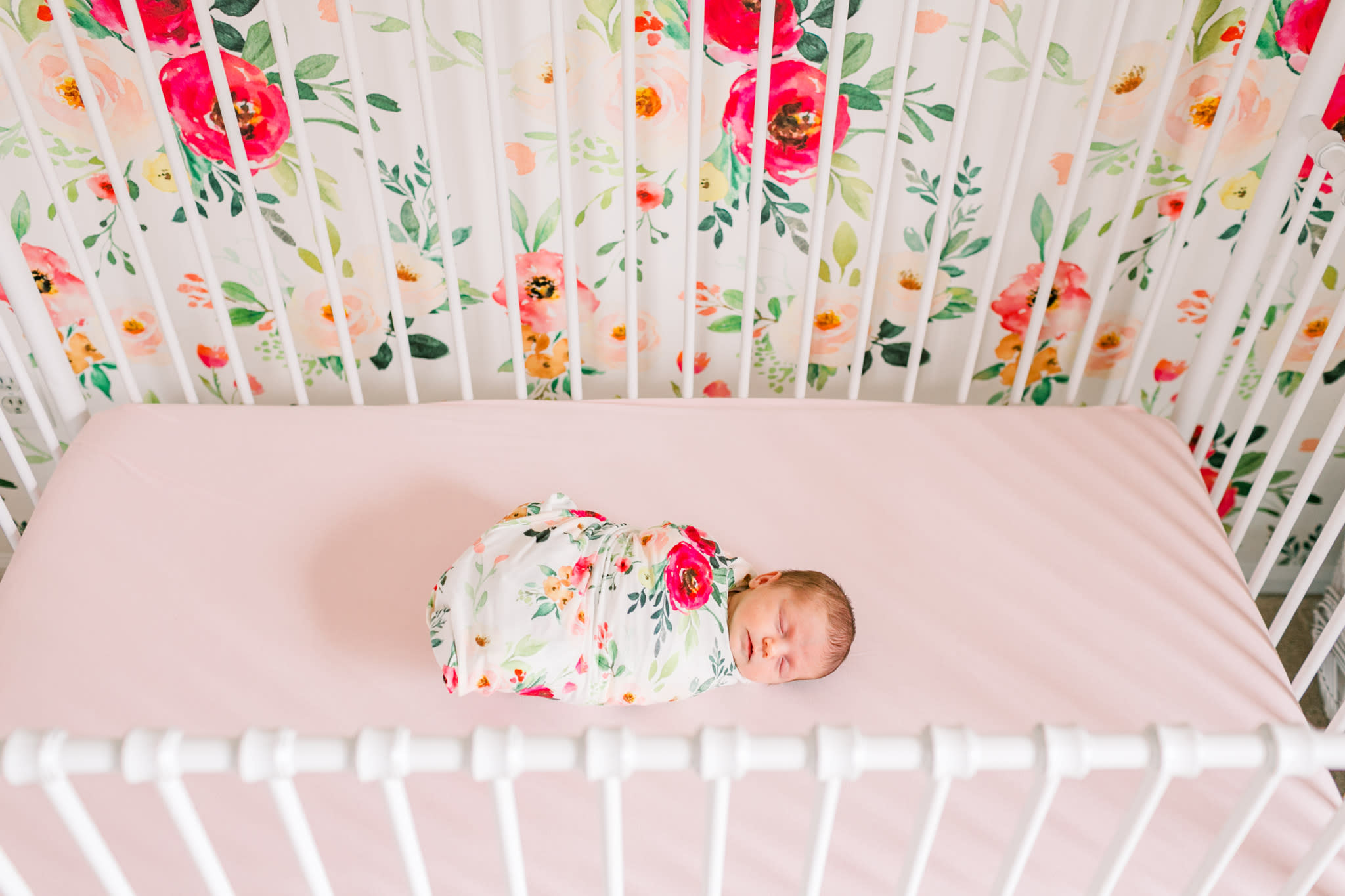 Baby swaddled in floral swaddle that matches the wallpaper behind the white crib she is laying in.