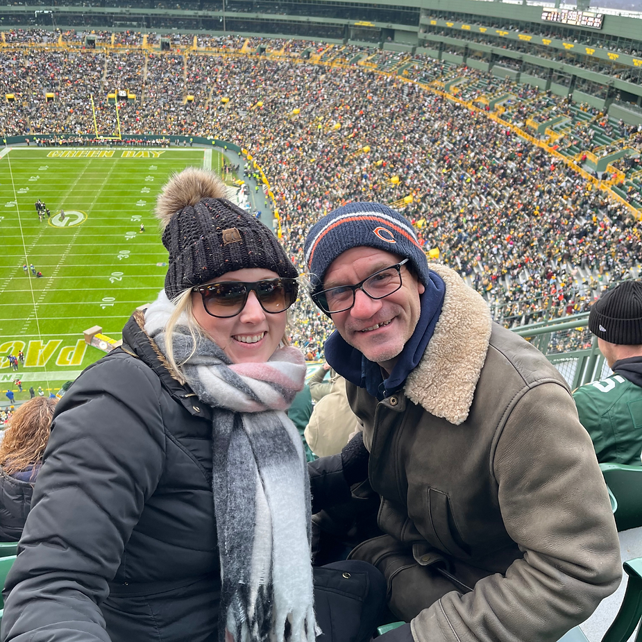Nicole and her husband in winter clothing smiling at a crowded Lambeau Field. Green field in background, text "Green Bay Packers" visible. Cold, festive atmosphere.
