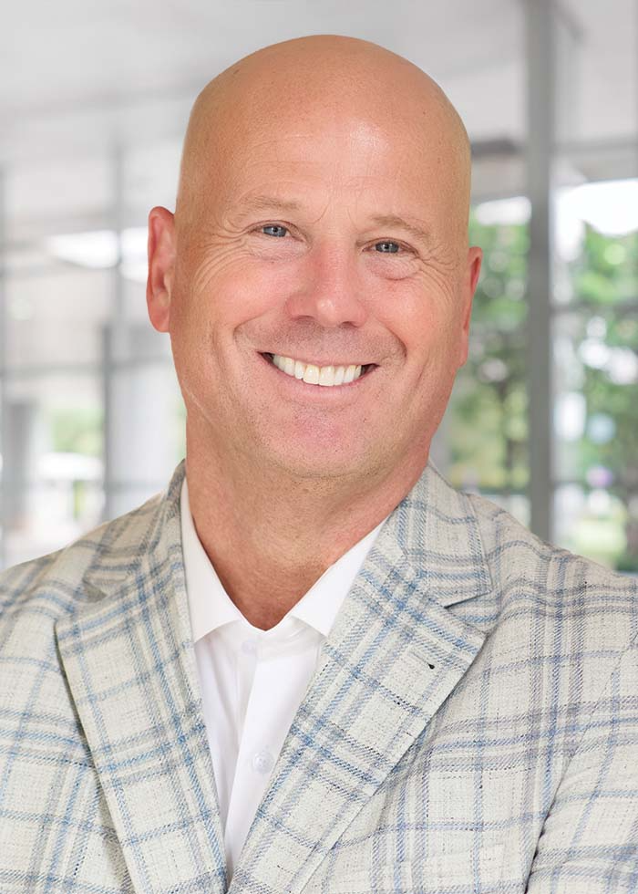 Smiling man in a light plaid jacket and white shirt, standing in a bright, modern indoor setting with windows.