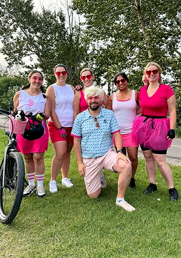 Group of five women and one male standing and smiling wearing pink and white Barbie and Ken costumes in summer outdoor setting with bicycle