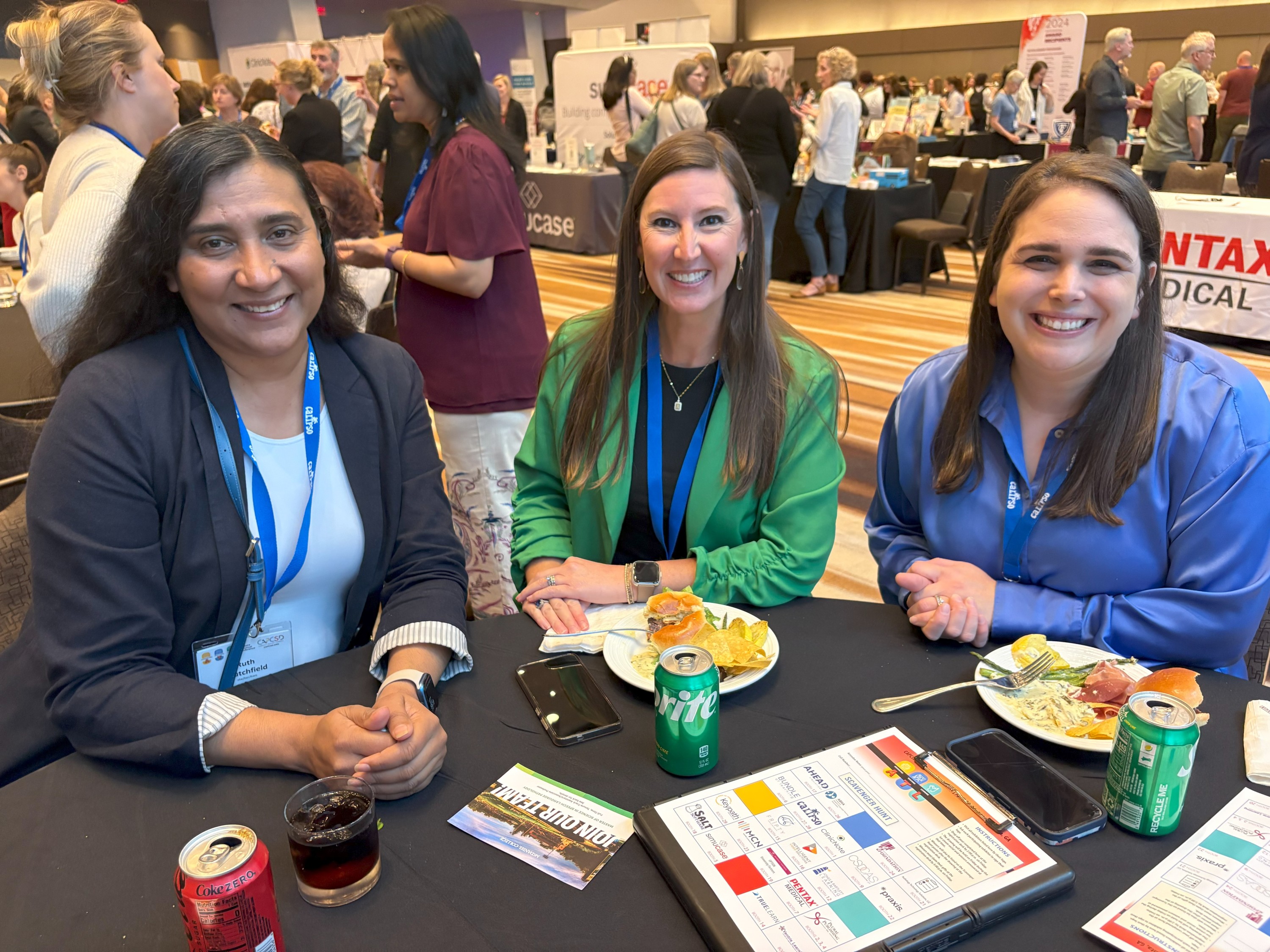 Three women wearing conference lanyards sitting at table with lunch plates and conference scavenger hunt cards