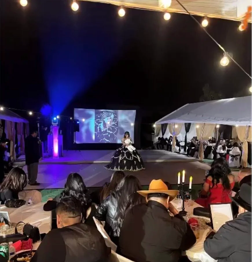 Nighttime outdoor event featuring a woman in a black and white dress dancing on a stage, surrounded by seated guests at decorated tables, with a large screen and string lights overhead.