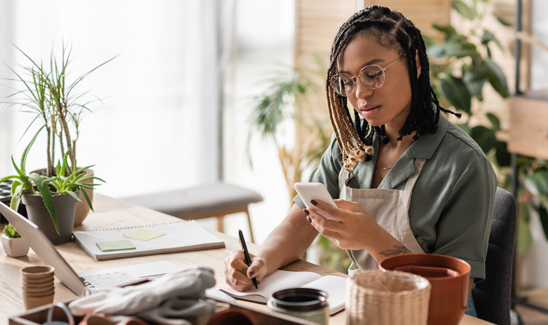 Focused female small business owner looking at smartphone and taking notes at desk