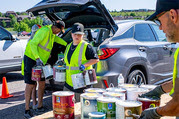 Several people in reflective vests carrying cans of paint from the trunk of a car to a table
