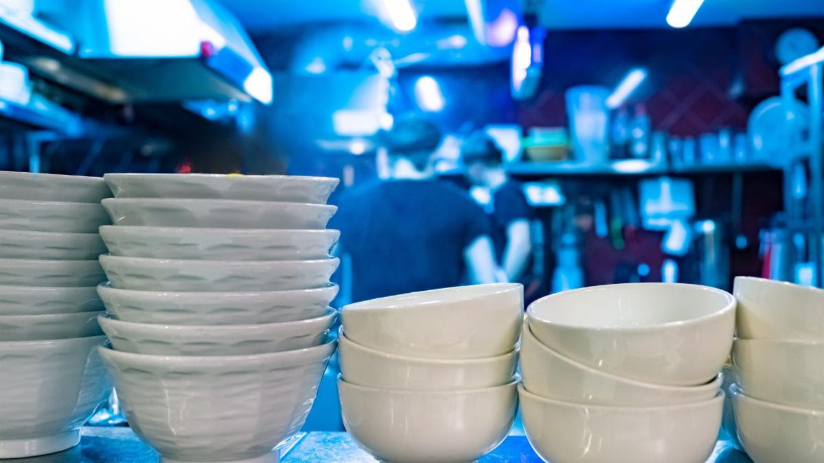 Stacks of reusable dishware on a shelf with a blurry restaurant scene in the background