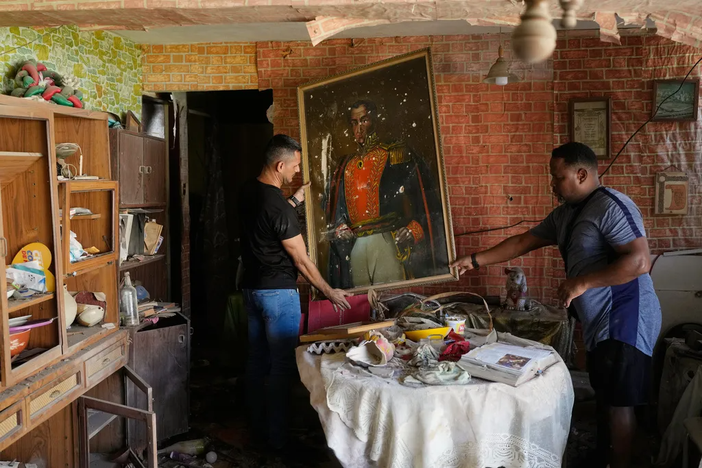 Two men stand in a cluttered, damaged room, inspecting a large portrait of a military figure while debris is scattered on the table and floor.