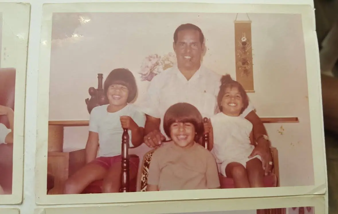A man sits on a chair with three smiling children, two girls and one boy, posing indoors in front of a wall with a hanging decoration and a flower arrangement.