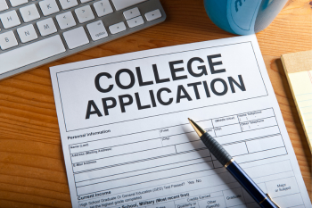 A college application form on a wooden desk next to a keyboard, pen, and a blue cup.