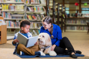 A woman and a boy read a book to a dog in a cozy library setting, surrounded by shelves of books.