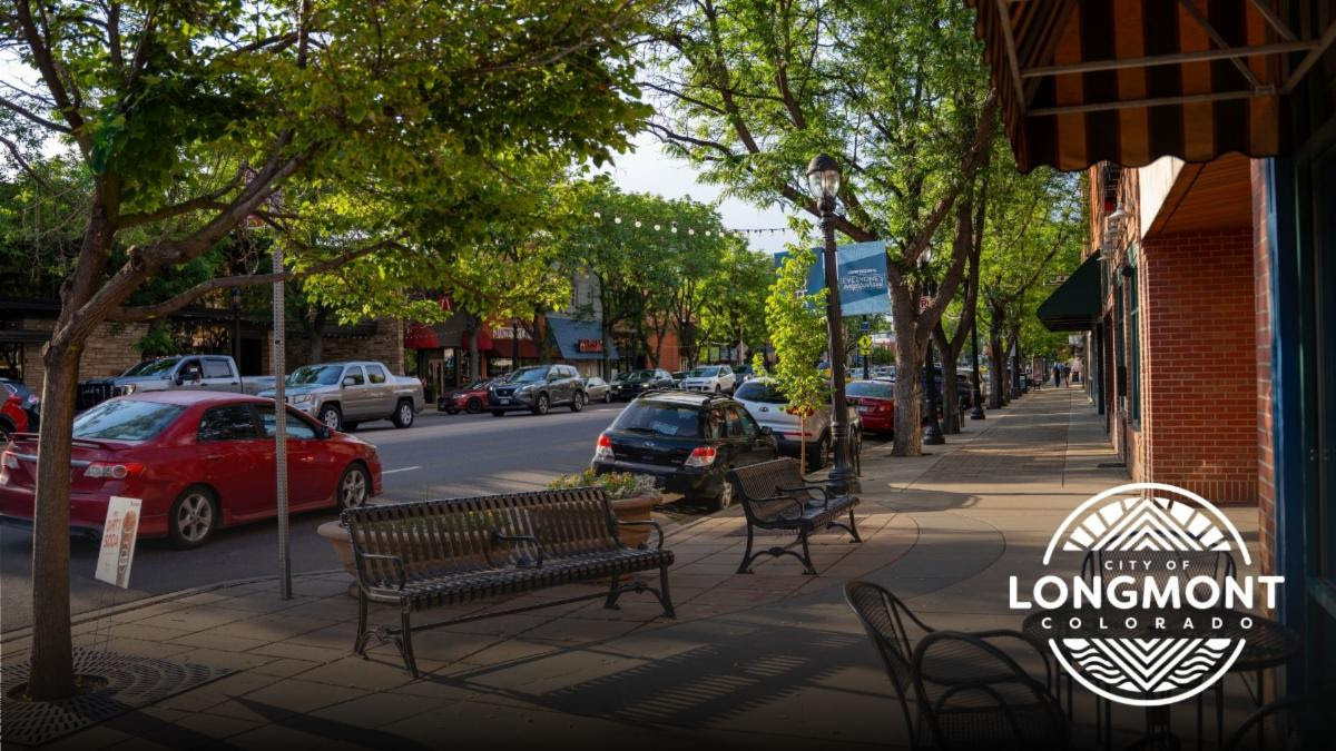 A scene from downtown Longmont shows businesses and restaurant. Cars drive by and an empty bench is inviting visitors. The City of Longmont logo is also shown.