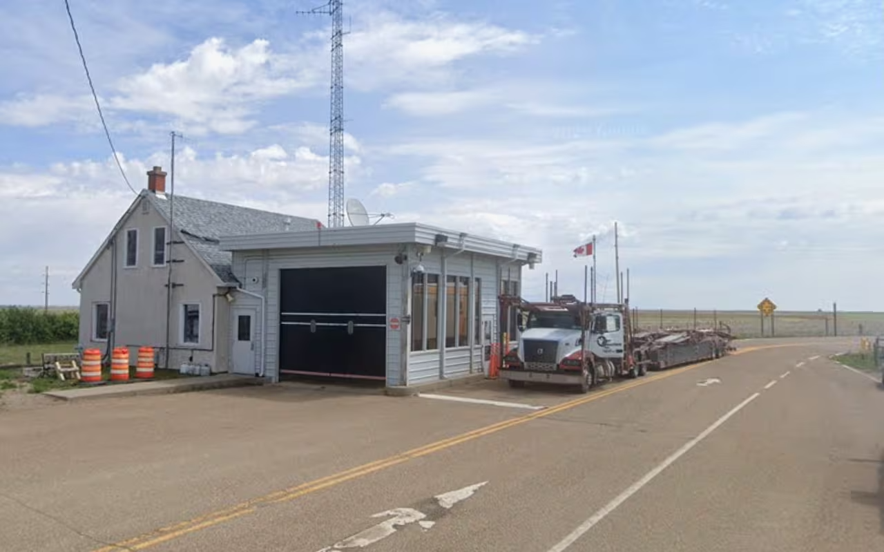 An aging farm house and attached garage sits on a highway with wide stretches of prairie grass in the background.