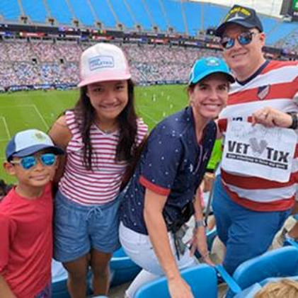 Family of four stand in stadium seating overlooking a football field, with one person holding a sign that reads “VET TIX” as a crowd fills the background.