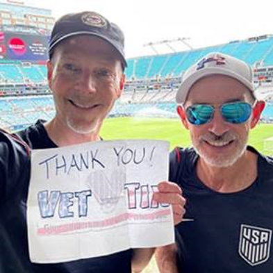 Two people standing in stadium seating hold a handwritten sign that reads, “Thank you! Vet Tix — Give Something Back to Those Who Serve,” with a football field visible in the background.