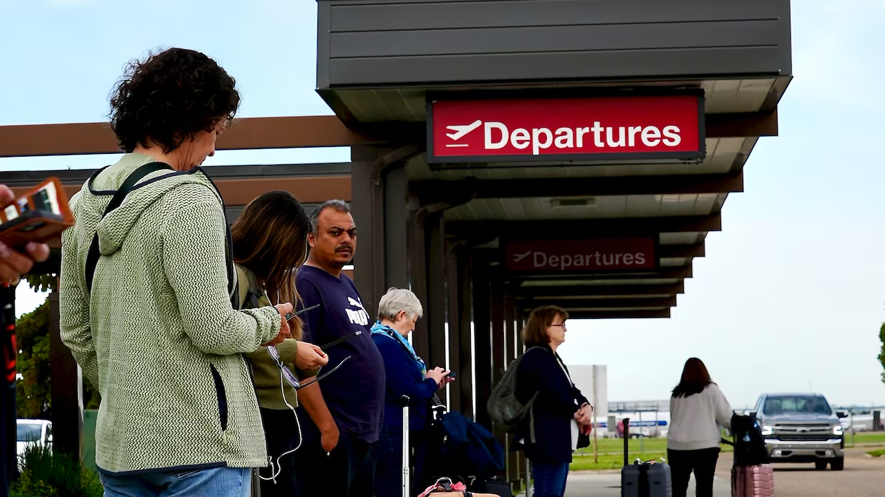 A line of passengers await for rides at a curb outside an airport terminal.