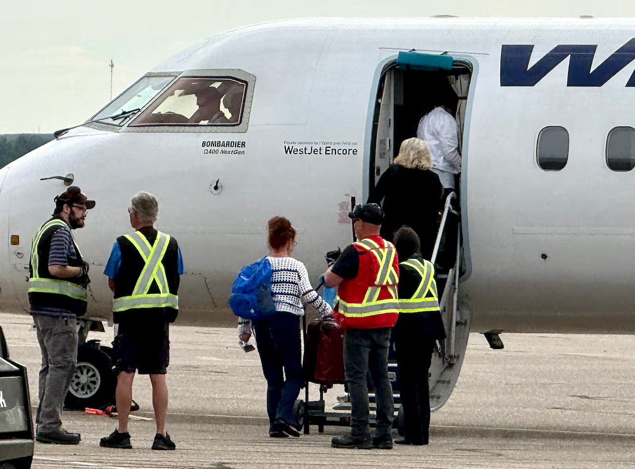 Airplane travellers wheel luggage to the steps of a plane waiting on a tarmac as crew stand nearby in reflective vests.
