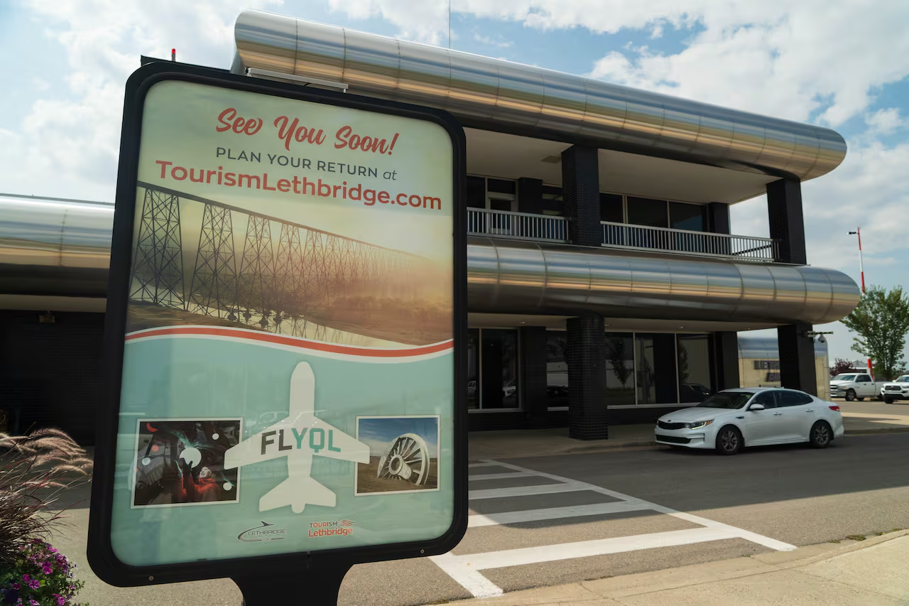 A large poster advertises tourism to Lethbridge in front of an airport terminal with stylized metal awnings.