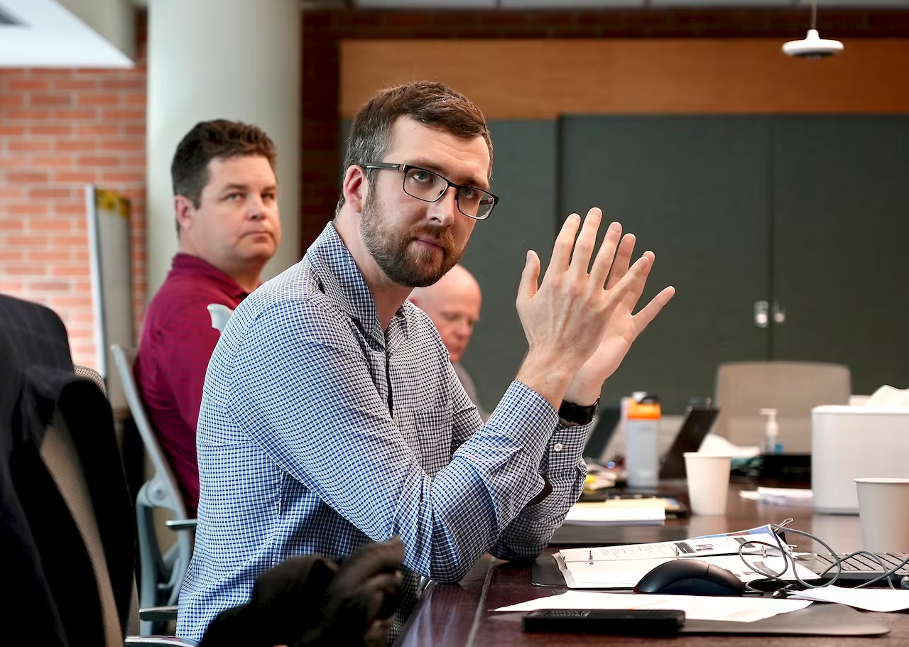 A man in a checkered shirt and glasses concentrates on a presentation (off camera) while gesturing with his hands.