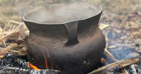 A traditional ceramic pot with a dark, rustic finish sitting amidst a small outdoor fire. The pot features interwoven patterns near the top and has a white, slightly flared rim. Flames and embers surround the pot. 