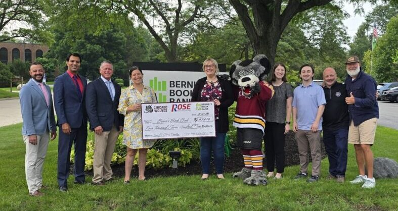 A group of people, including a mascot, stand outside on grass holding a large check. Trees and parked cars are in the background near a sign that reads “Bern Boo.”.