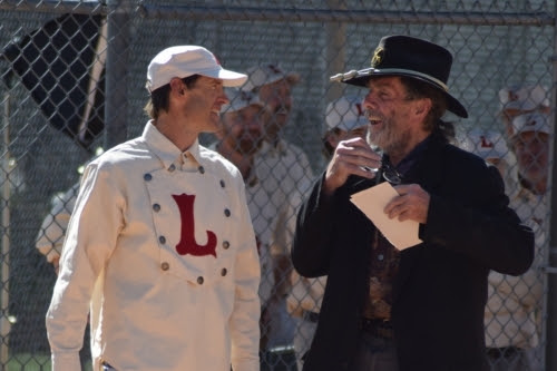 A photo of a man wearing a vintage baseball uniform with his head turned to look at another man in a western style hat and jacket.