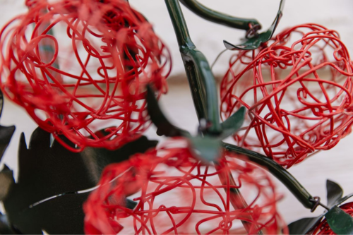 A close-up photo of a wire sculpture depicting three red tomatoes on a green leafy vine.