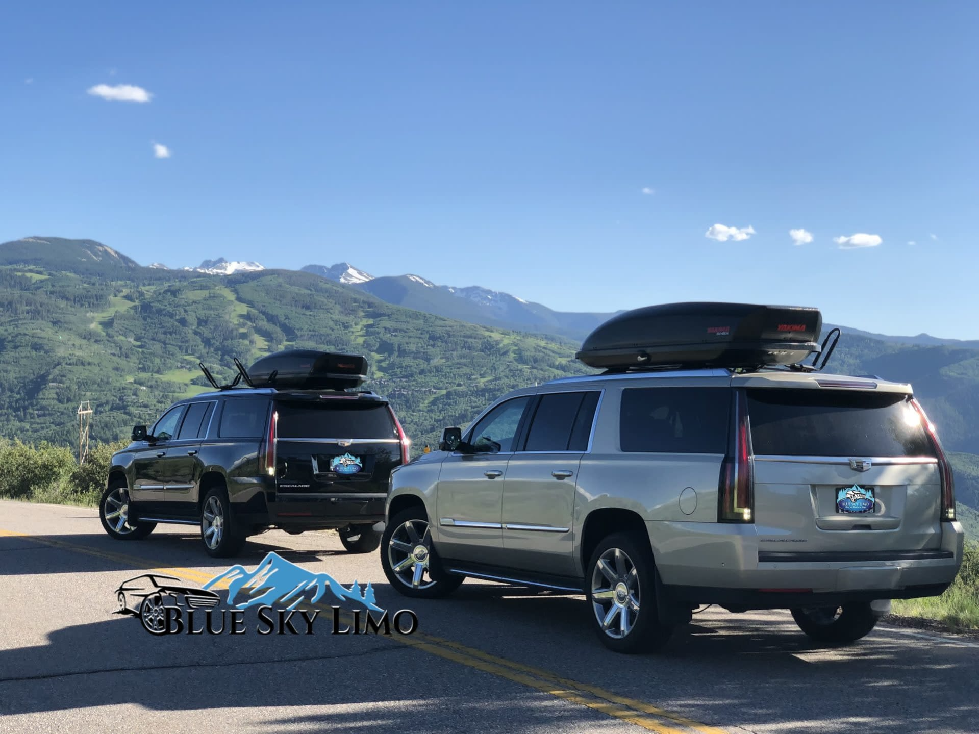 Two Blue Sky Limo luxury SUVs with rooftop cargo boxes against a mountain backdrop