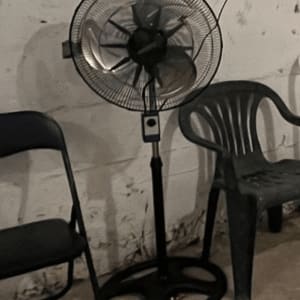 מאוורר עומד ליד כיסאות פלסטיק וקיר בטון