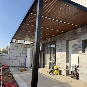 פרגולת אלומיניום שחורה עם שלבי הצללה דמוי עץ ותאורת לד מובנית בחצר