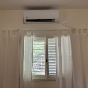 מזגן מותקן על קיר מעל חלון עם וילון לבן