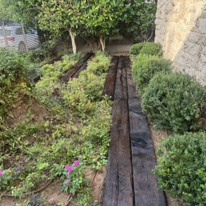 שביל קורות עץ מאסיביות מעוצב בגינה לצד צמחייה וקיר אבן
