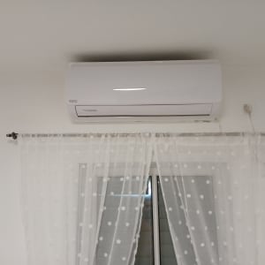 מזגן מותקן על קיר מעל חלון עם וילון לבן