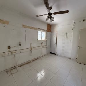 חדר ריק עם מאוורר תקרה, קירות חשופים ורצפת אריחים לבנה.