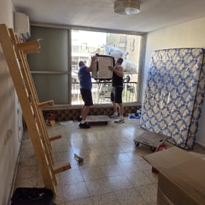 שני אנשים מעבירים רהיטים בחדר ריק עם מזרן וקרטונים