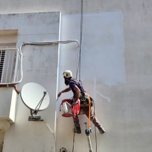 אדם צובע קיר חיצוני בעזרת רתמה ודלי צבע