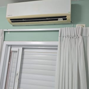 מזגן מותקן על קיר מעל חלון עם וילון לבן