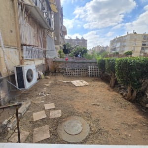 חצר עם אדמה יבשה, שיחים ומבנים ברקע