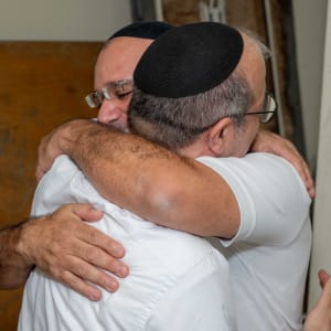 שני אנשים מתחבקים, חובשים כיפות, ברקע קיר עץ.